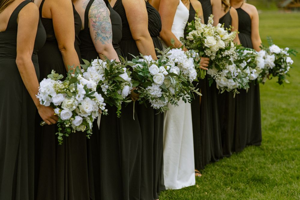 Bridesmaids in black gowns holding white floral bouquets stand beside bride in white dress on green lawn.