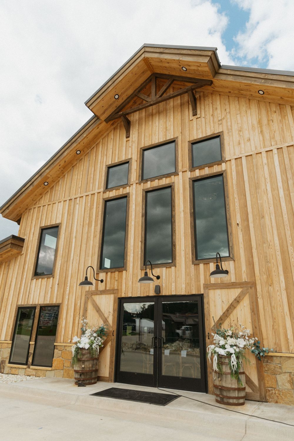 Rustic wooden barn wedding venue exterior with large windows, barn doors, and floral barrel decorations.