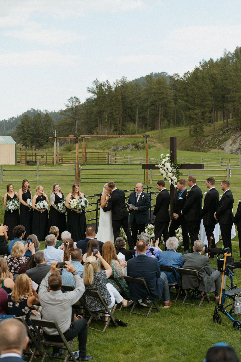 Outdoor wedding ceremony on green meadow with bridal party in black, guests seated, pine trees and hills in background.