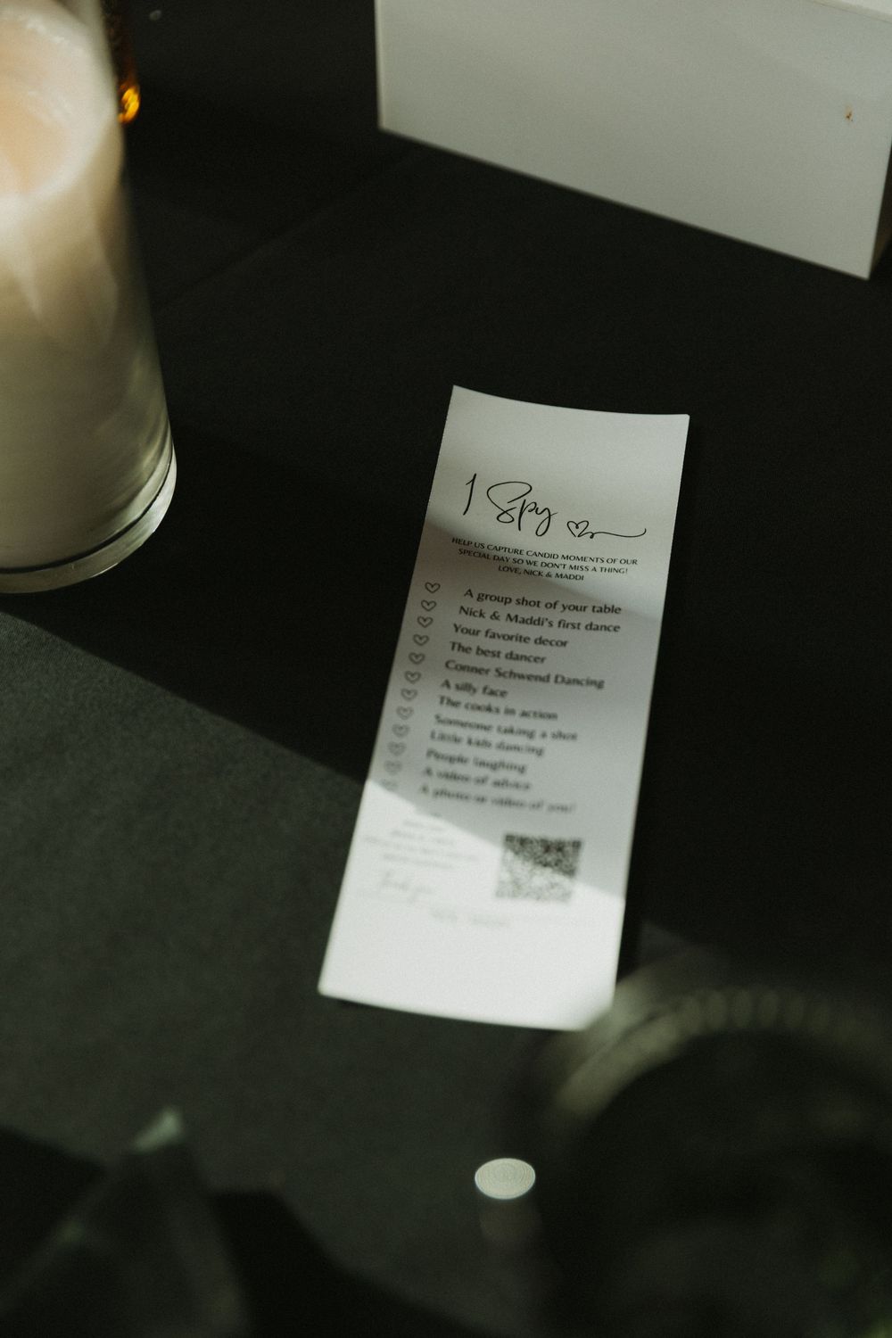 Wedding reception bingo card on black table next to white candle in glass holder.