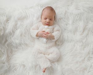 Newborn baby sleeping peacefully on white fluffy rug wearing cream knit romper suit.
