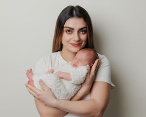 Young mother in white t-shirt tenderly holds sleeping newborn baby wrapped in fluffy white blanket against neutral background.
