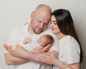 A couple in white shirts tenderly holds their sleeping newborn baby against a soft neutral background.