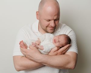 Bald father tenderly holds sleeping newborn baby in white outfit against neutral background.
