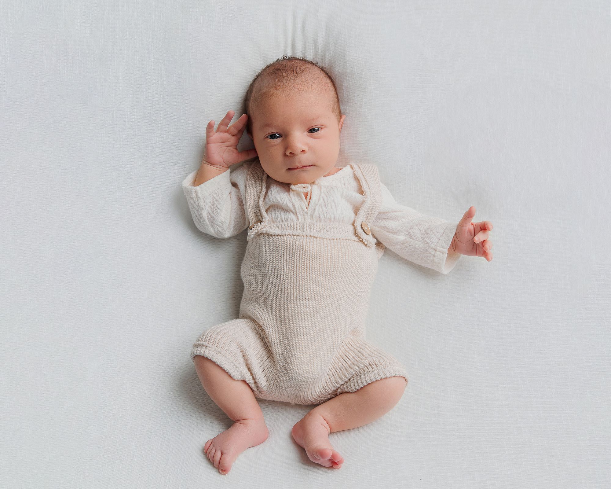 Newborn baby in cream knit romper lying on white background, peaceful infant portrait photography.