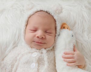 Smiling newborn baby in white fuzzy bear outfit holding a small toy on a fluffy white blanket.