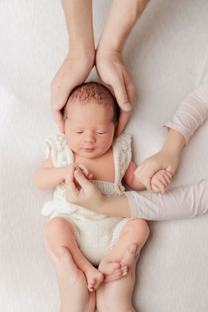 Newborn baby sleeping peacefully while parents gently cradle head and hold tiny hands and feet on white background.