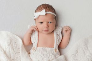 Newborn baby girl in white knit romper and bow headband lying on soft white blanket.