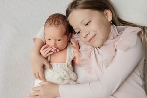 A young girl in a pink dress tenderly holds a sleeping newborn baby dressed in a white knit outfit.