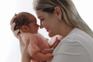 A young mother tenderly holds her newborn baby in a pink knit outfit, touching foreheads in an intimate moment.