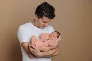 A father in a white t-shirt tenderly holds his sleeping newborn baby against a warm beige background.