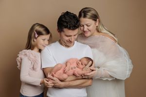 Family of four posing with newborn baby, parents and young daughter smiling warmly against beige backdrop.