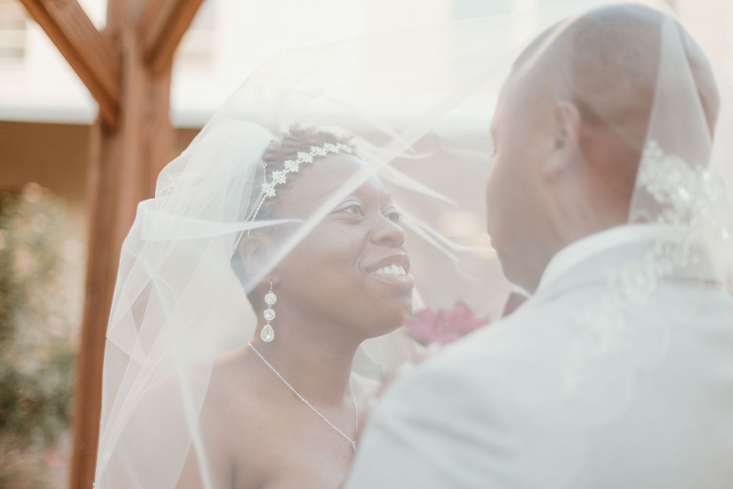 Beautiful bride smiling at groom under flowing white veil during outdoor wedding ceremony.