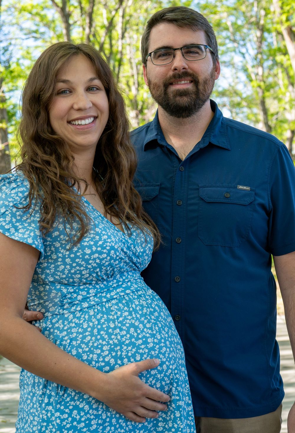 Smiling pregnant woman in blue floral dress poses with bearded man in navy shirt outdoors among trees.