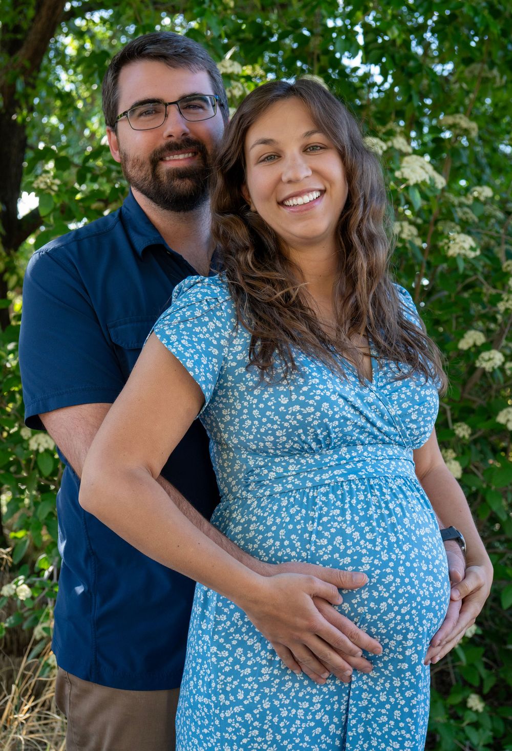 Smiling pregnant woman in blue floral dress with partner embracing her outdoors.