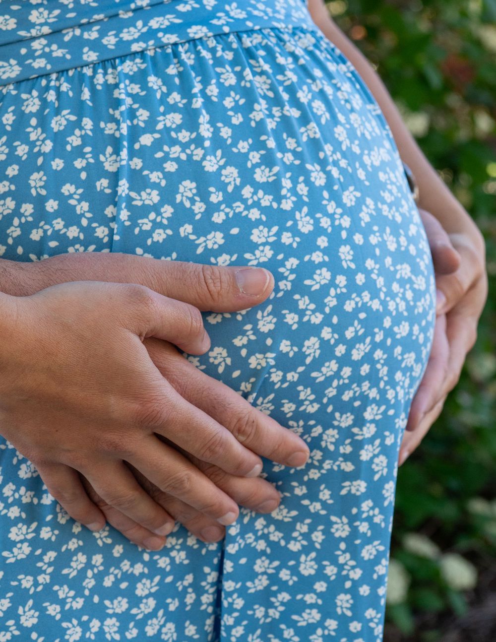 Close-up of pregnant woman in blue floral dress with hands gently cradling her baby bump outdoors.