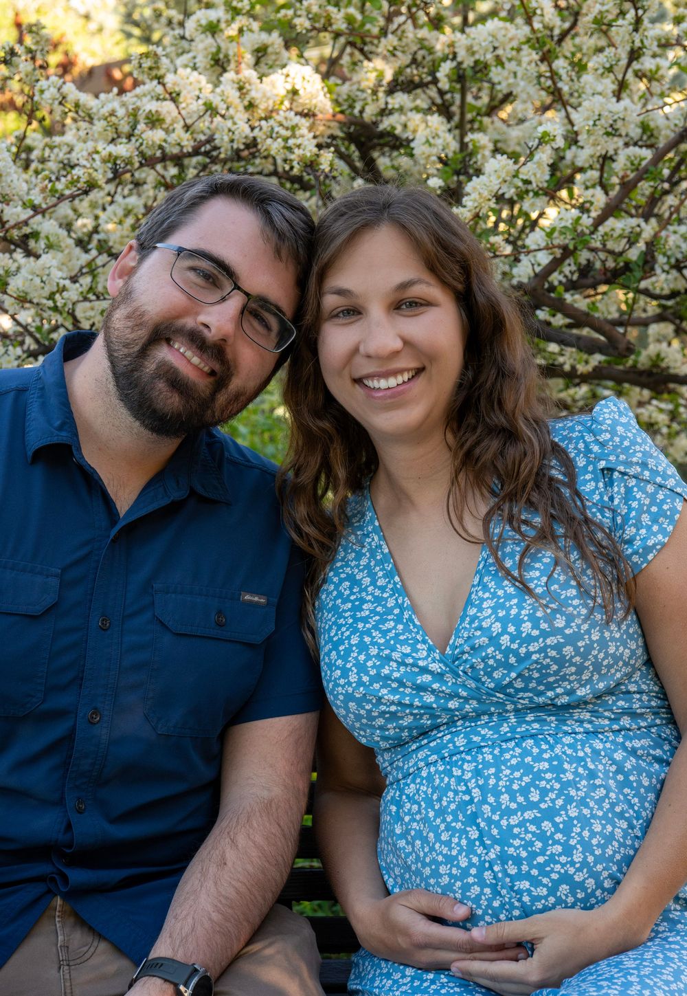 Smiling pregnant couple sitting together outdoors in front of a blooming white flower tree on a sunny day.