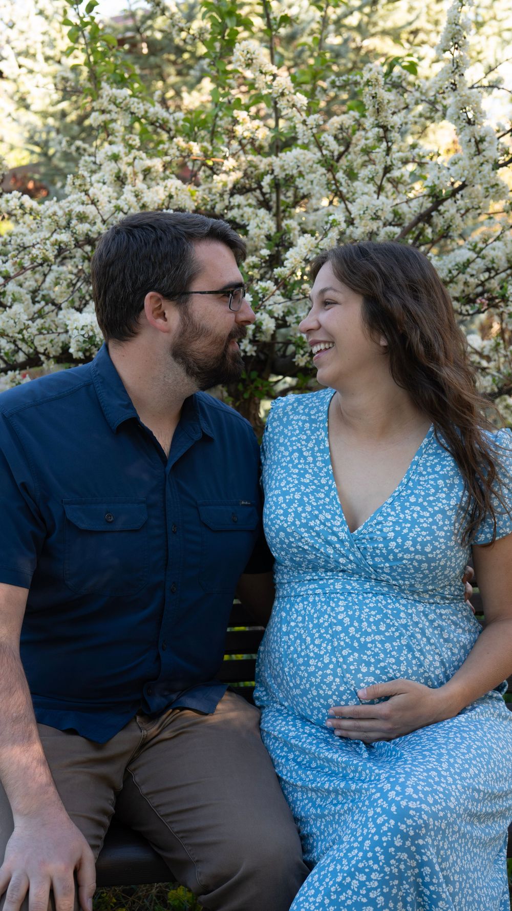 A smiling pregnant woman in a blue floral dress sits with her partner on a bench near a blooming white flowering tree.