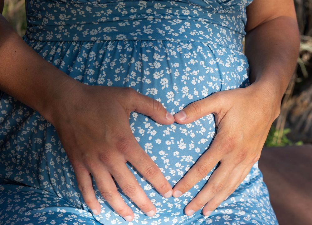 Pregnant woman in blue floral dress forming heart shape with hands on baby bump.