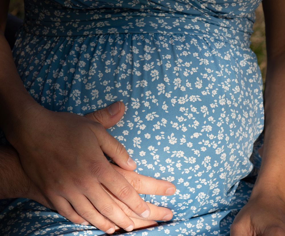 Pregnant woman in blue floral dress gently cradling her baby bump with both hands outdoors.