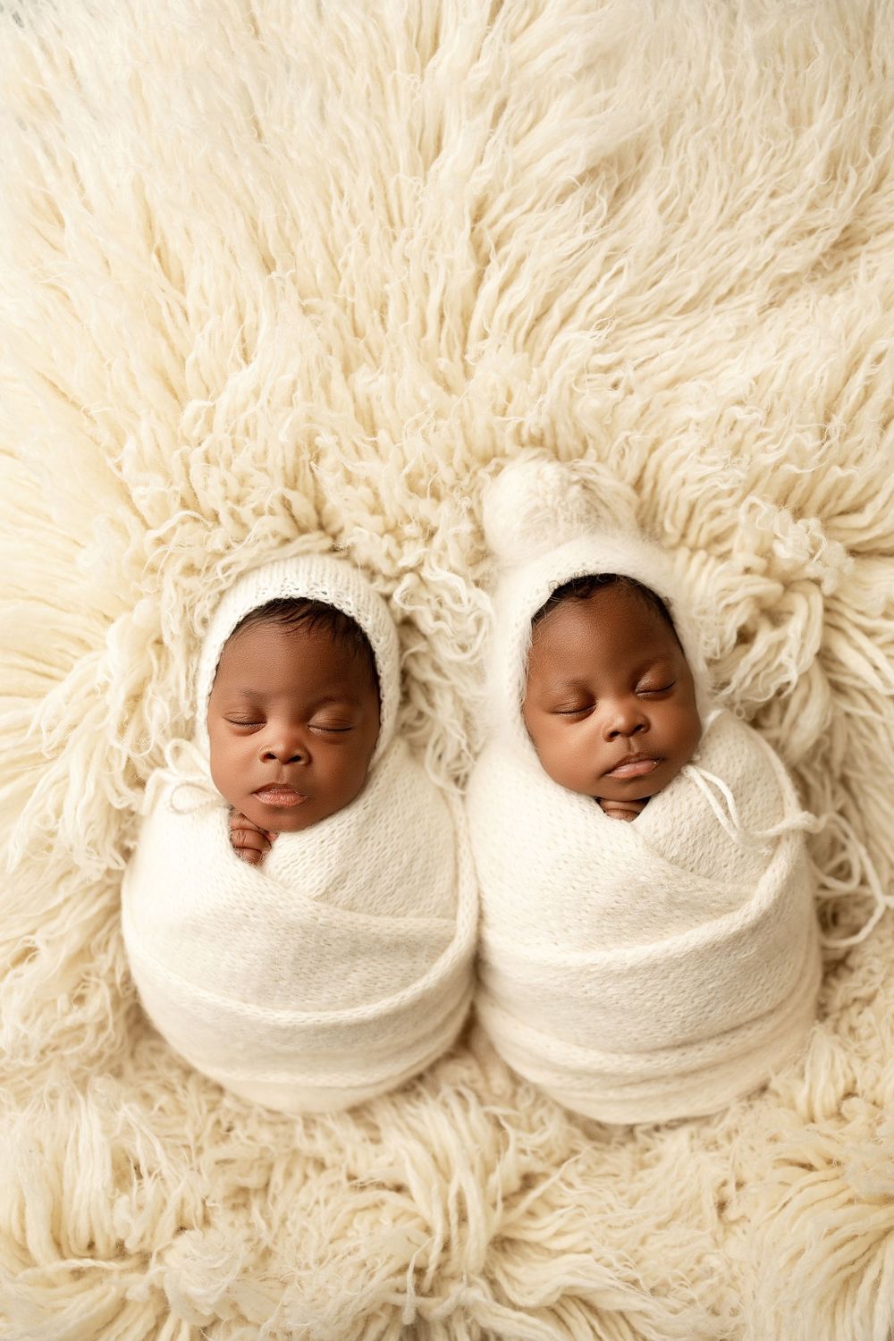 Two sleeping newborn twins swaddled in white wraps and bonnets lying side by side on a cream fluffy blanket.