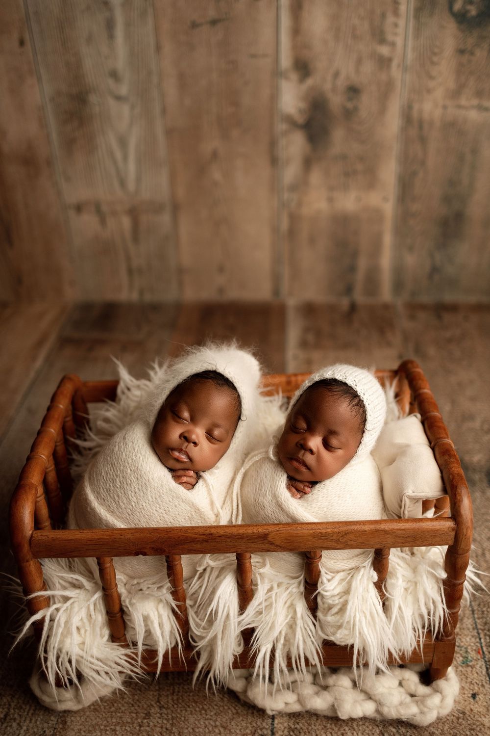 Two sleeping newborn twins wrapped in white swaddles wearing bonnets, nestled in a small wooden bed with fluffy white bedding.