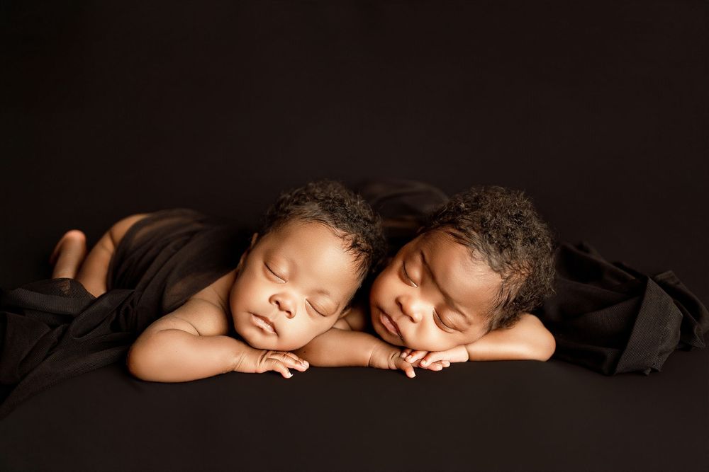 Two sleeping newborn twins lying side by side on a dark background, wrapped in soft dark fabric.