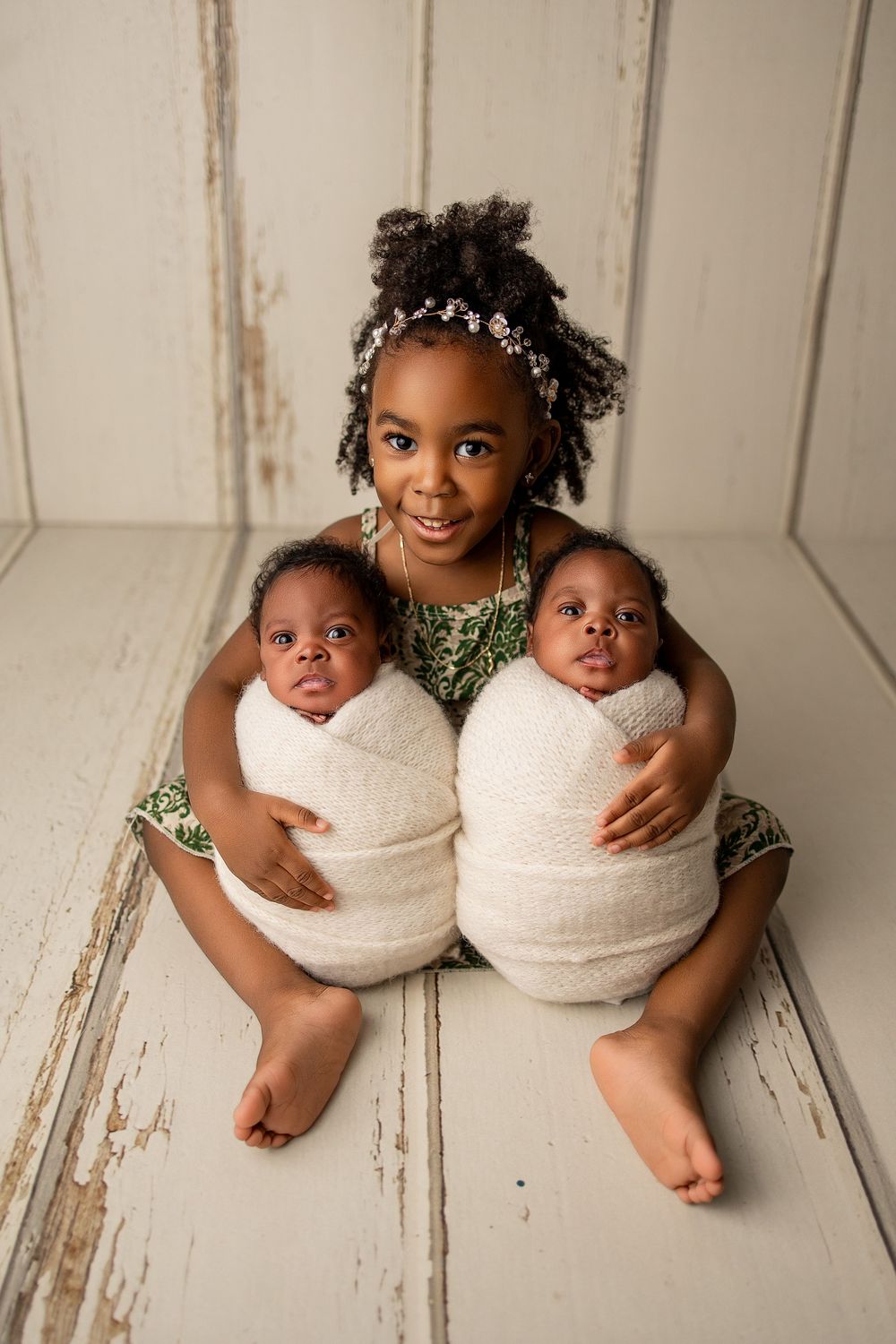 Young girl with floral headband holds two swaddled newborn twins while sitting on rustic white wooden floor.