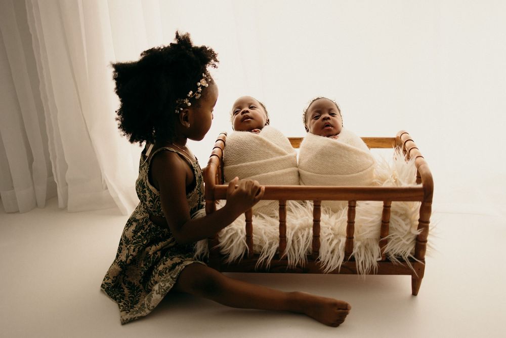 Young girl sits beside a wooden crib holding two swaddled newborn twins in a bright white studio setting.
