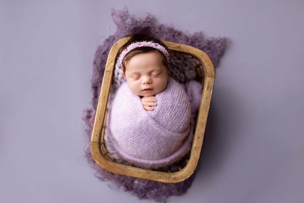 Newborn baby swaddled in lavender wrap sleeping peacefully in wooden bowl with purple fluffy blanket.