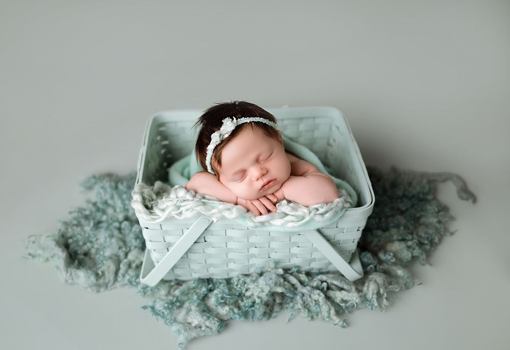 Sleeping newborn baby girl in mint green outfit and floral headband posed in a white basket with soft teal floral layers.
