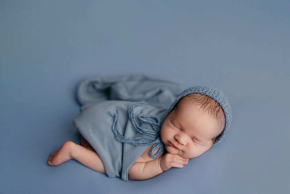 Sleeping newborn baby wrapped in soft blue knit fabric and bonnet, posed on a blue backdrop.