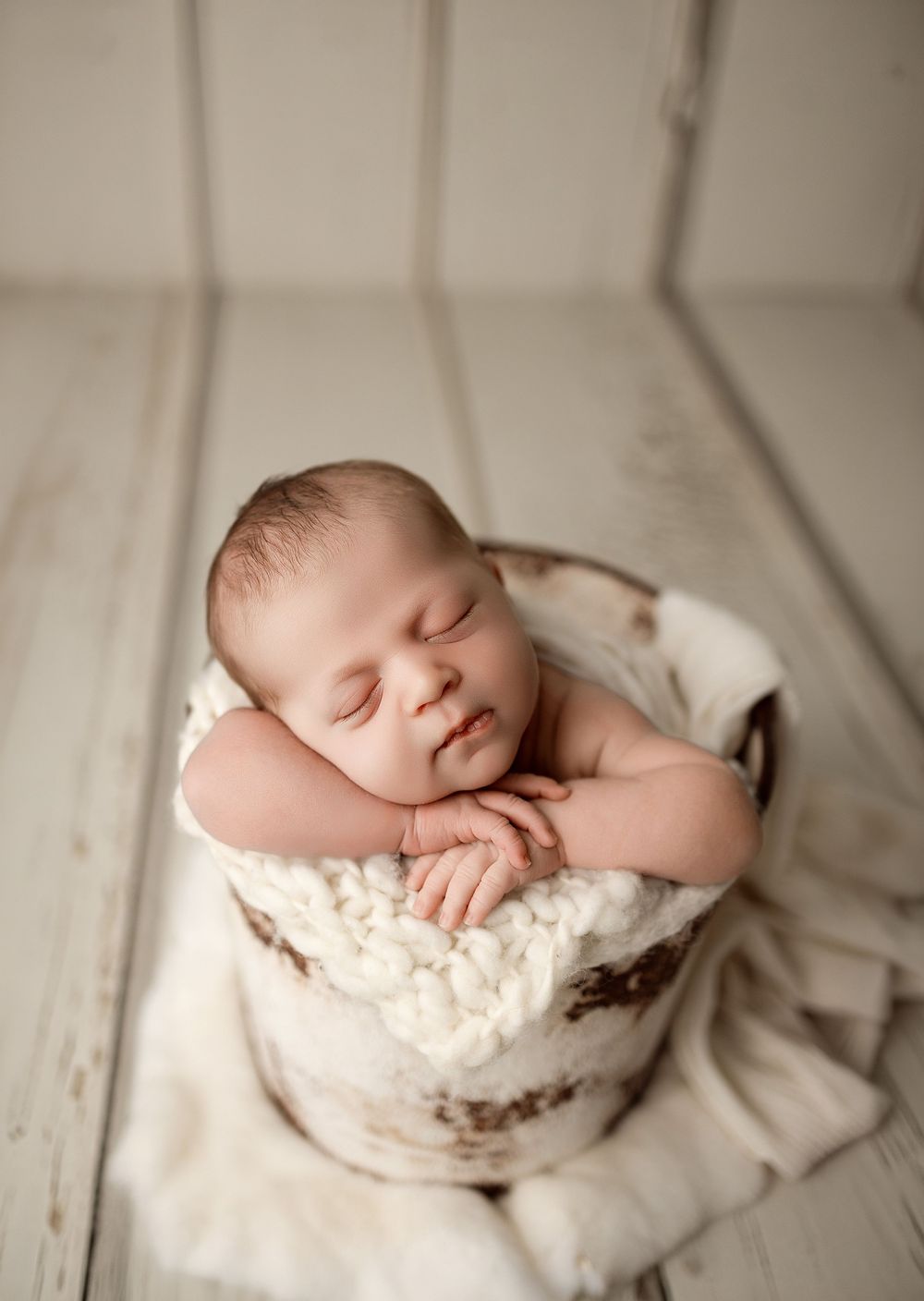 Sleeping newborn baby posed in a rustic wooden bucket with soft white knit blanket in a professional studio portrait.