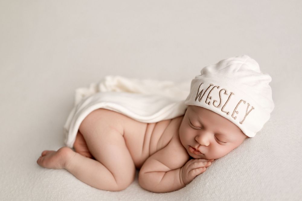 Sleeping newborn baby named Wesley wearing a white personalized hospital hat, curled up on a soft white blanket.