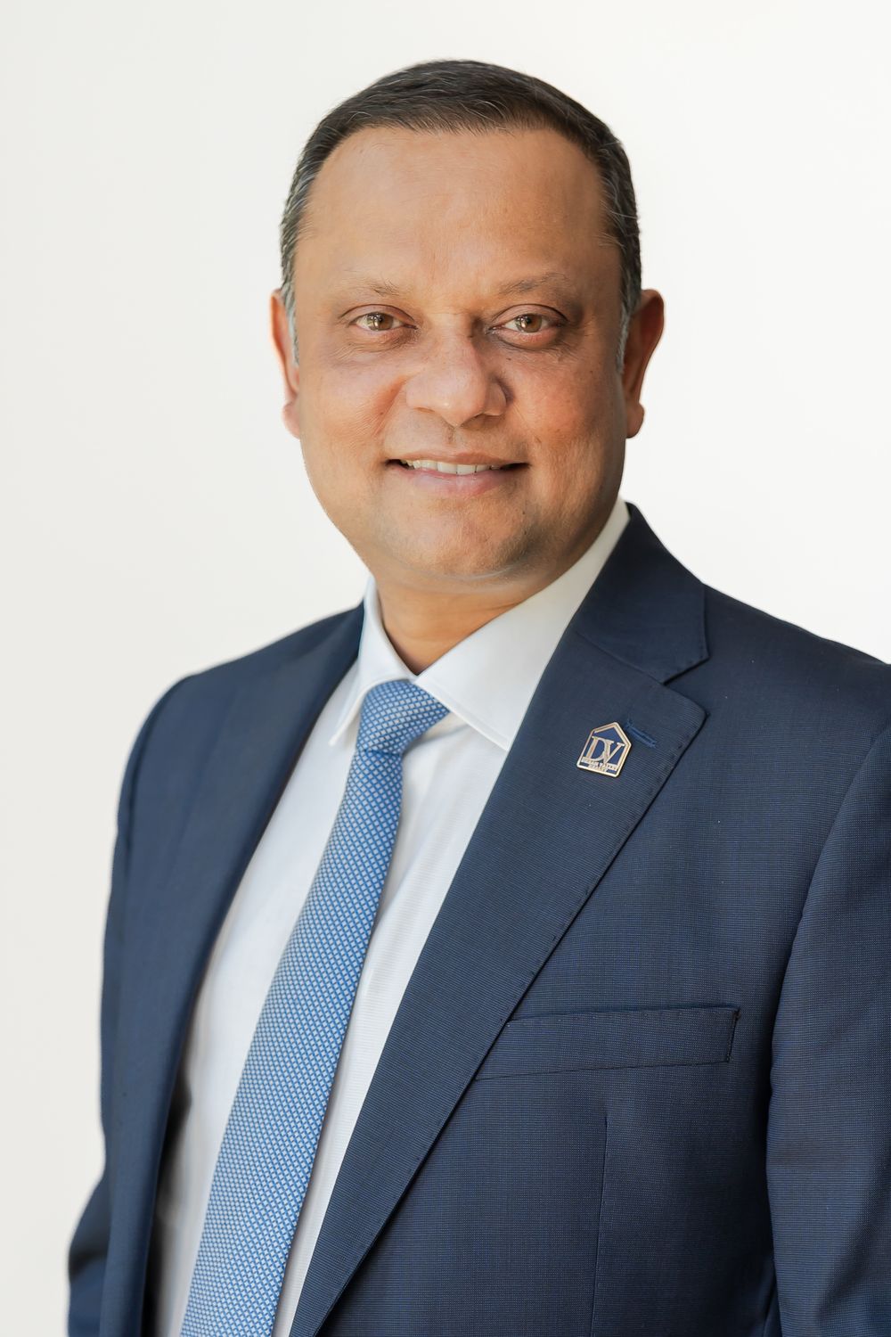 Professional man in navy suit and blue tie smiling confidently against a white background.