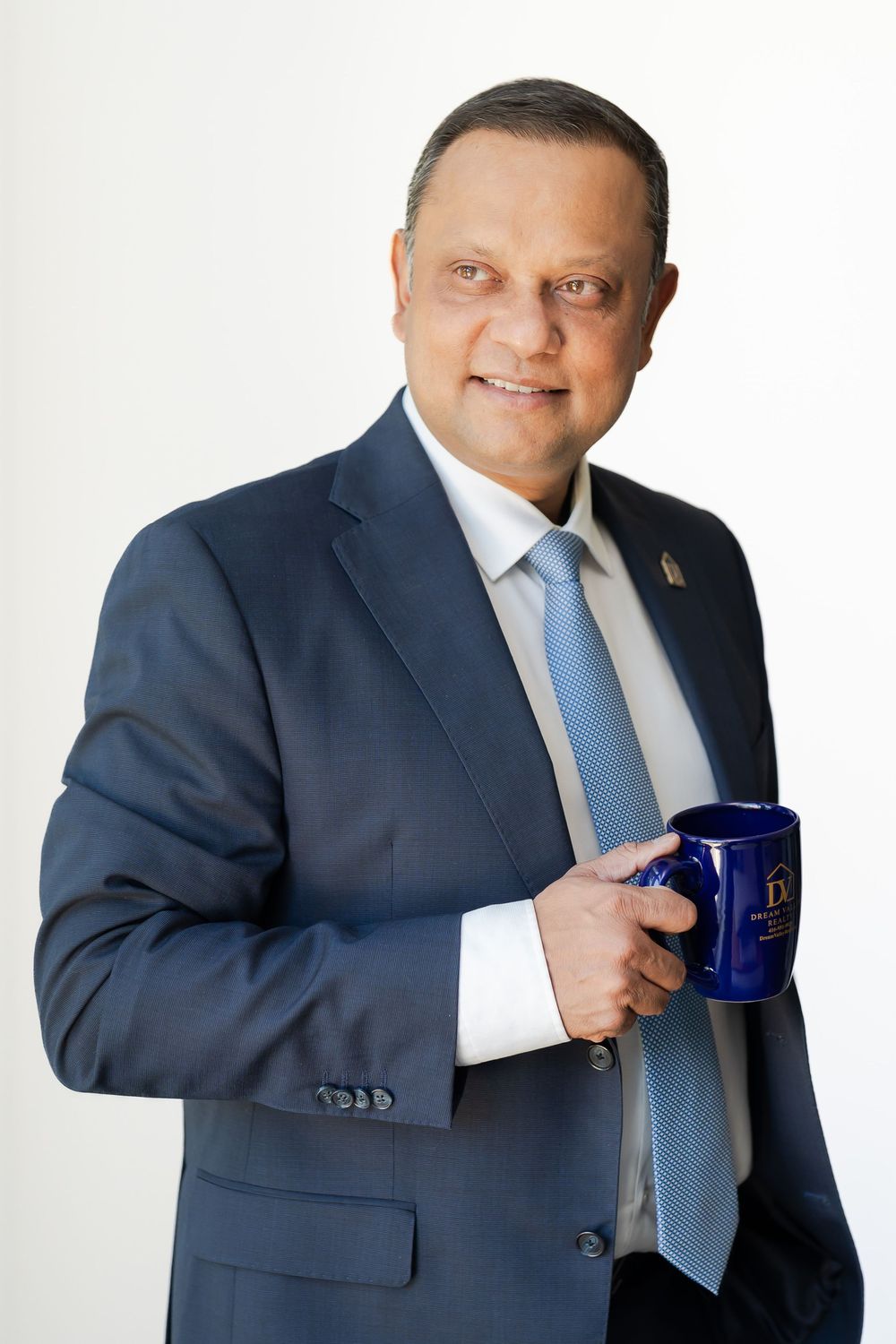 Professional man in blue suit holding a navy mug, smiling confidently against a white background.
