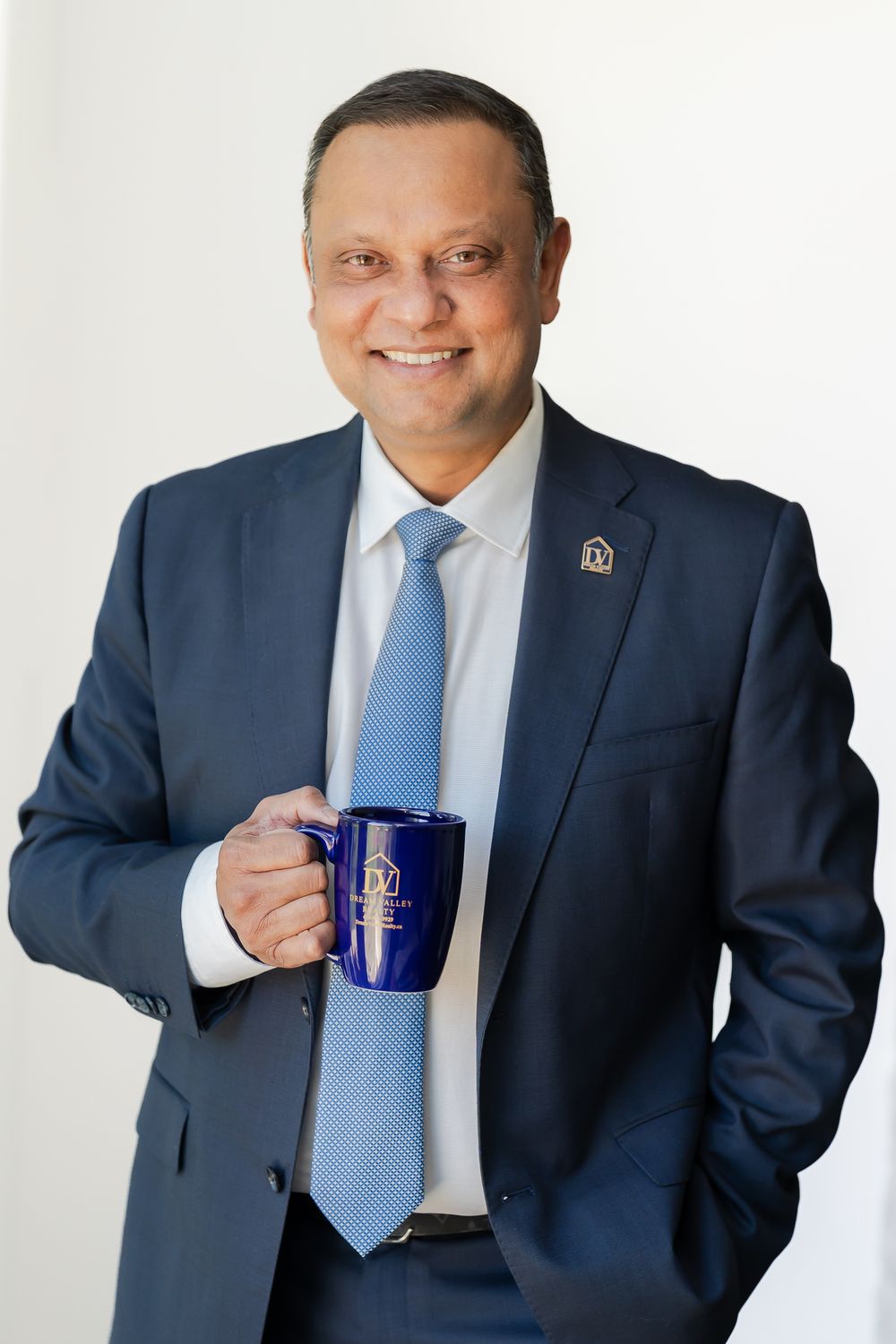 Professional businessman in navy suit holding a blue branded mug, smiling confidently against a white background.