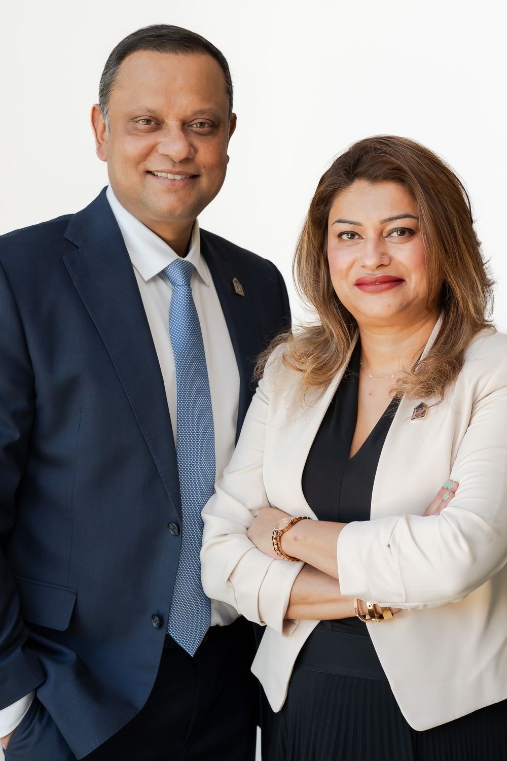 Professional portrait of a man in a navy suit and a woman in a beige blazer smiling confidently against a white background.