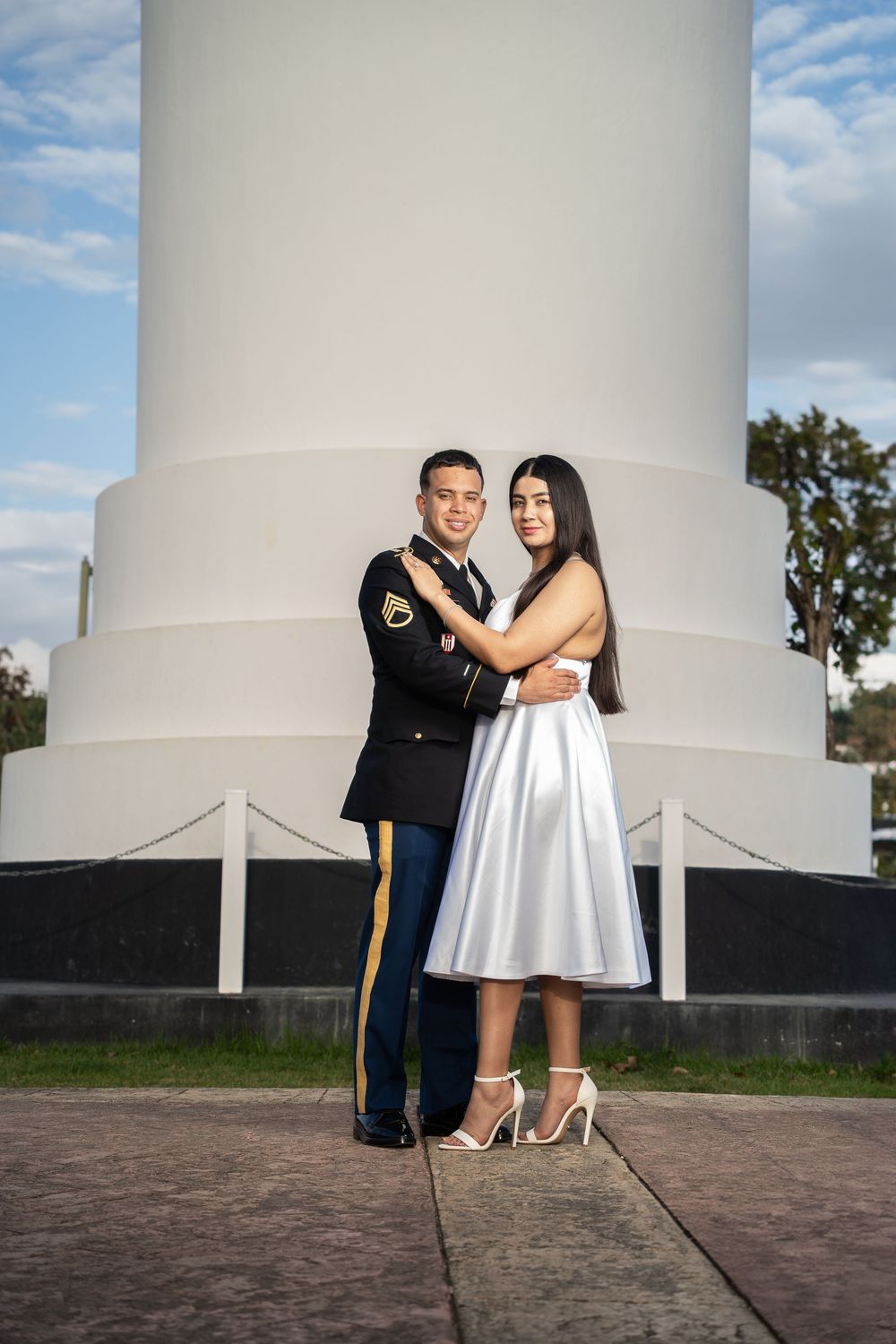 A soldier in dress uniform embraces a woman in a white dress near a monument outdoors.
