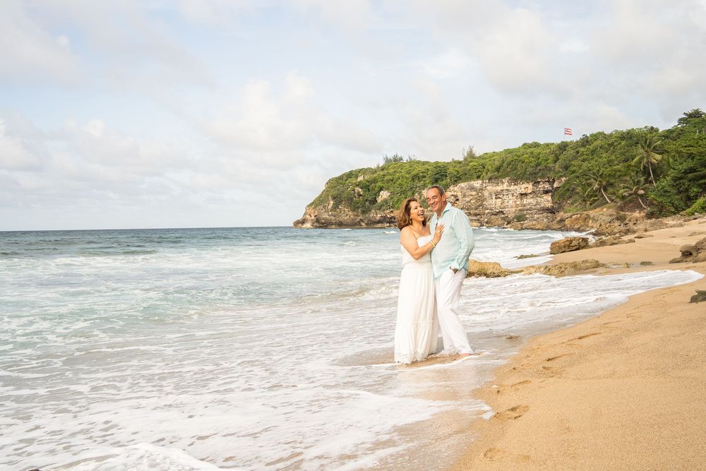 Couple embracing on a sandy tropical beach with ocean waves and lush green cliffs in the background.