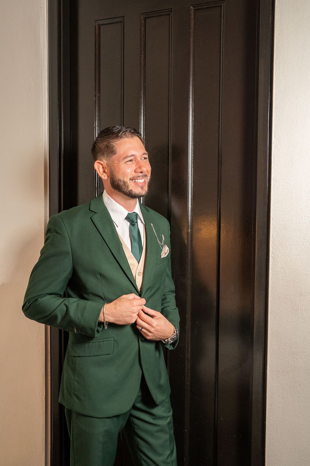 Smiling man in a green suit with matching tie and pocket square, standing by a dark door.