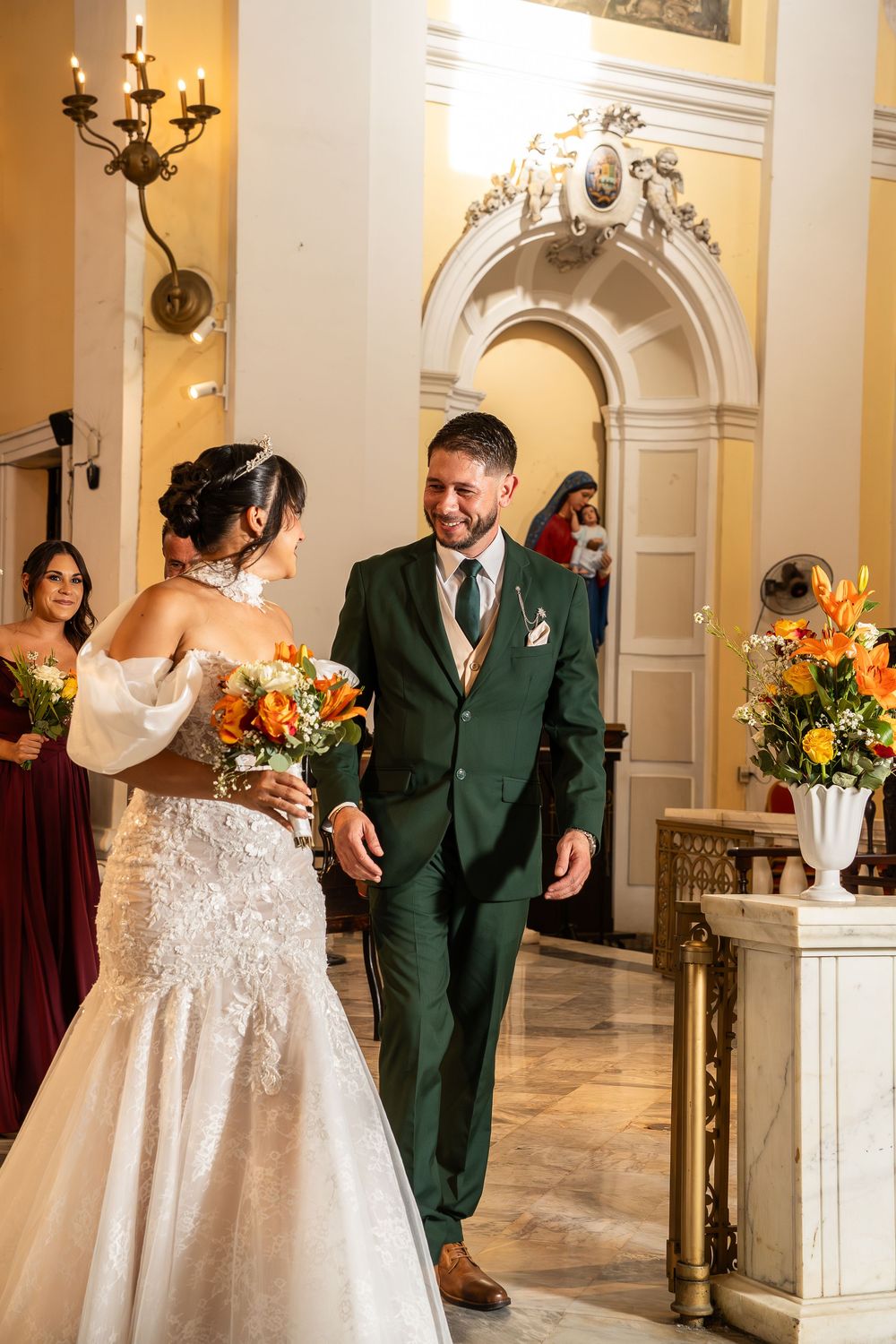 Bride and groom share a joyful moment at church altar, she in lace gown holding orange bouquet, he in green suit.