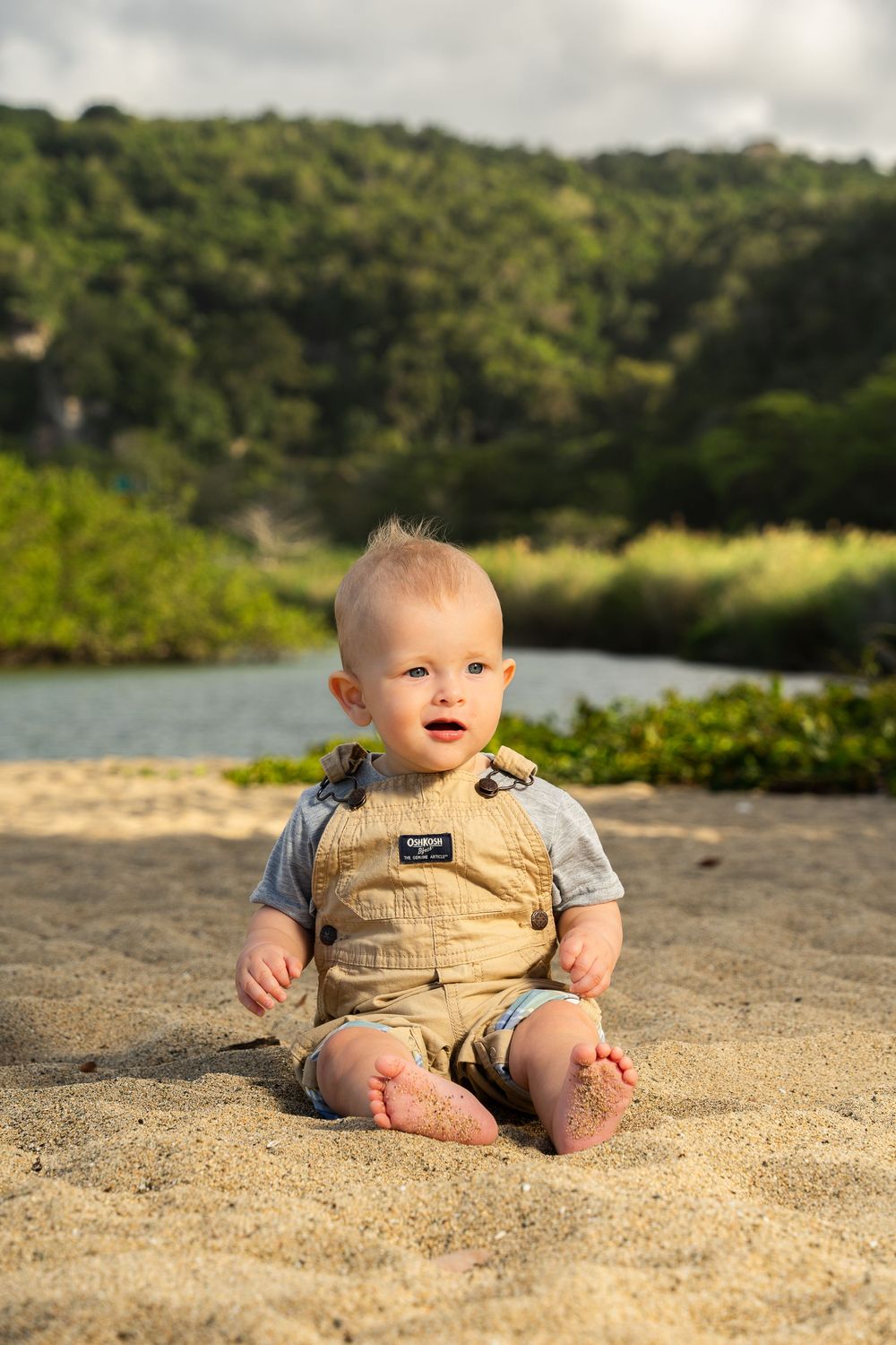 Baby boy in overalls sitting on sandy beach with lush green hills and calm water in the background.