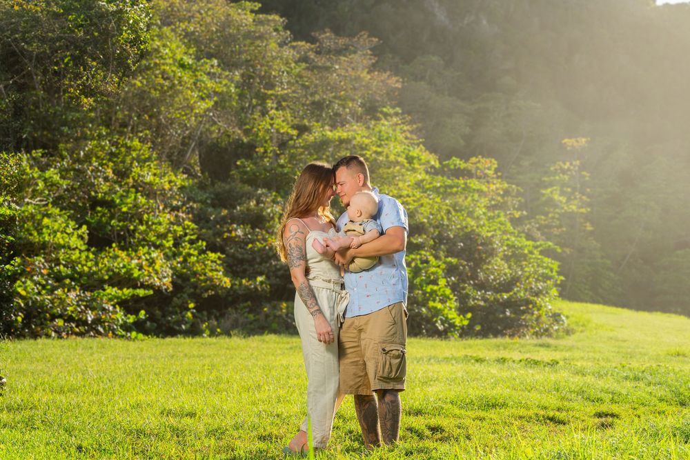 A couple shares a tender moment kissing while holding their baby in a sunlit green meadow surrounded by lush trees.