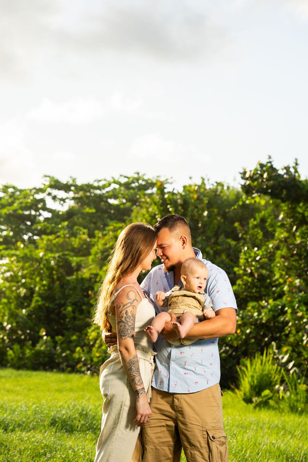Happy couple touching foreheads while holding infant baby outdoors in sunny green park setting.