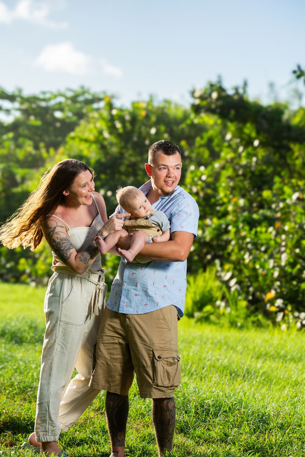 Young family with tattooed mother and father holding baby outdoors on green grass with lush trees in background.