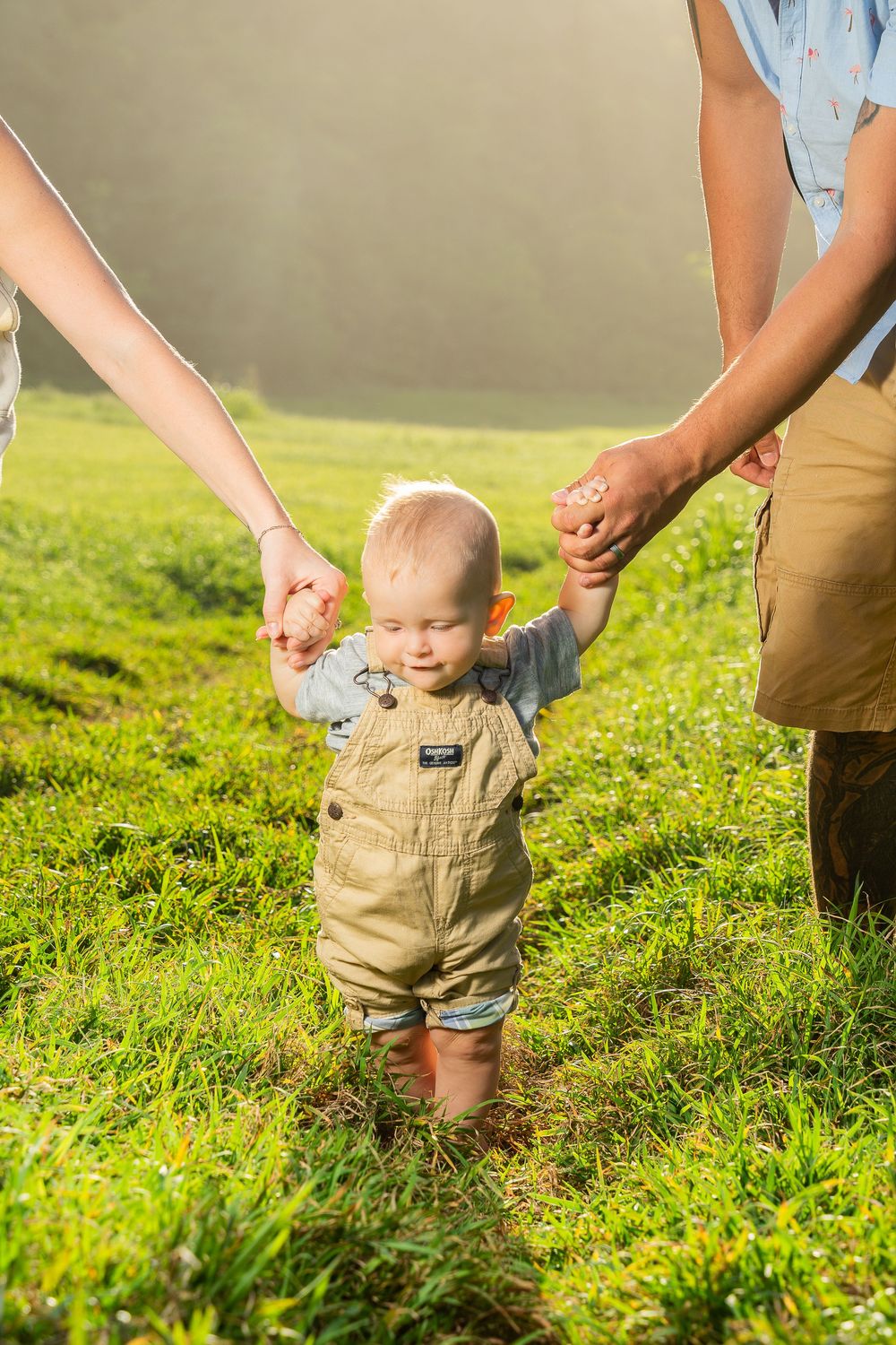 A toddler in overalls takes first steps on green grass, holding hands with two adults on a sunny day.