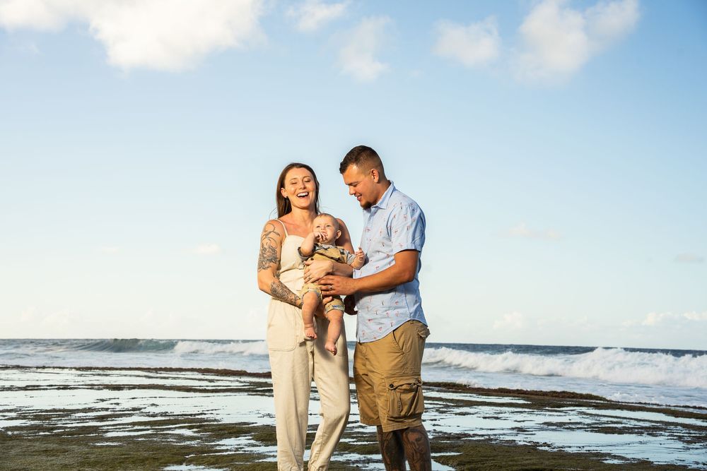 A happy couple stands on a beach holding their small dog, with ocean waves and blue sky behind them.