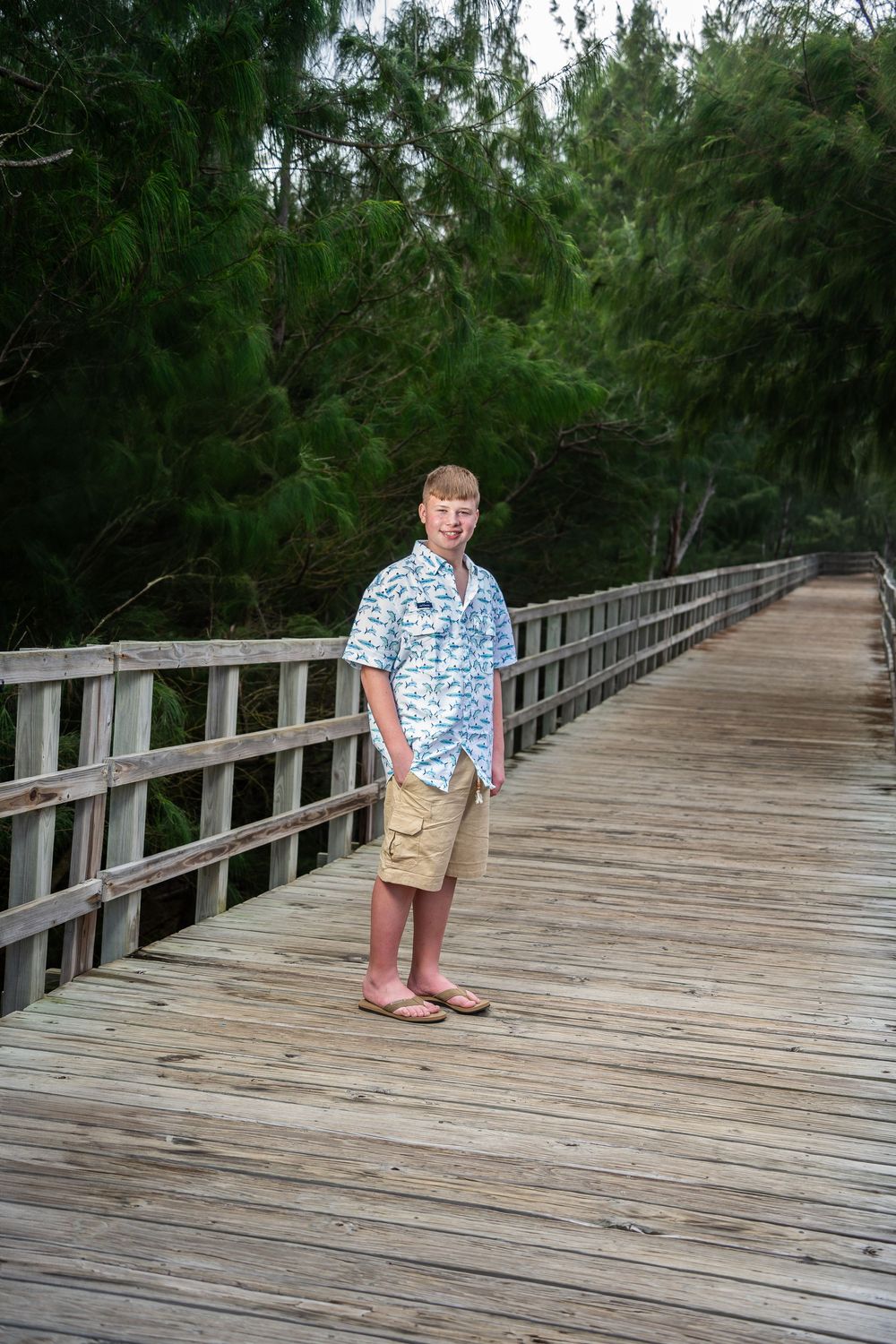 Young man in floral shirt and khaki shorts standing on a wooden boardwalk surrounded by lush green trees.