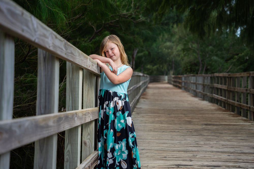 Young girl in teal top and floral skirt posing on a wooden bridge surrounded by lush green trees.
